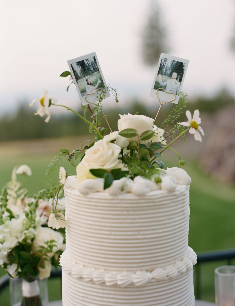 Two-tier white wedding cake with floral decoration and two Polaroid photos on top, set outdoors.