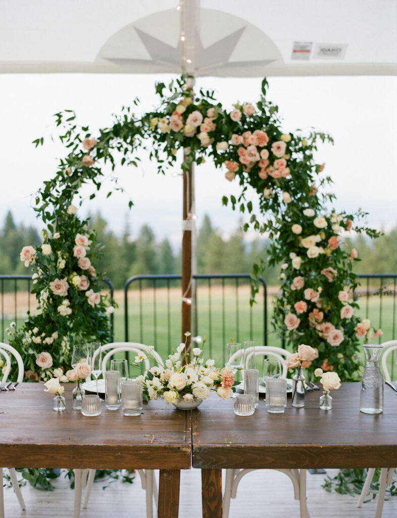 A table with floral arrangements is set under an arch made of greenery and pink roses, against a background of trees and fog.