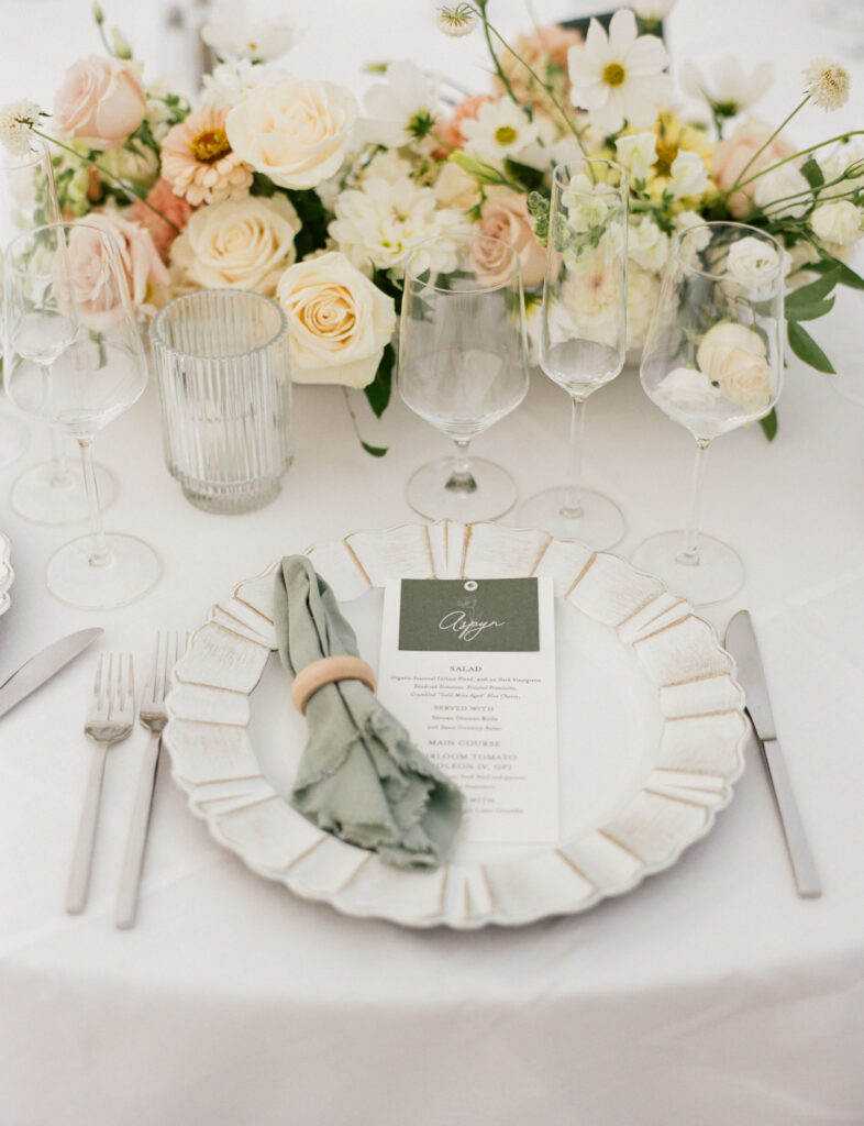 Elegant table setting with a white plate, a menu, a folded napkin, and silver cutlery. Three wine glasses are arranged on the table, accompanied by a floral centerpiece.