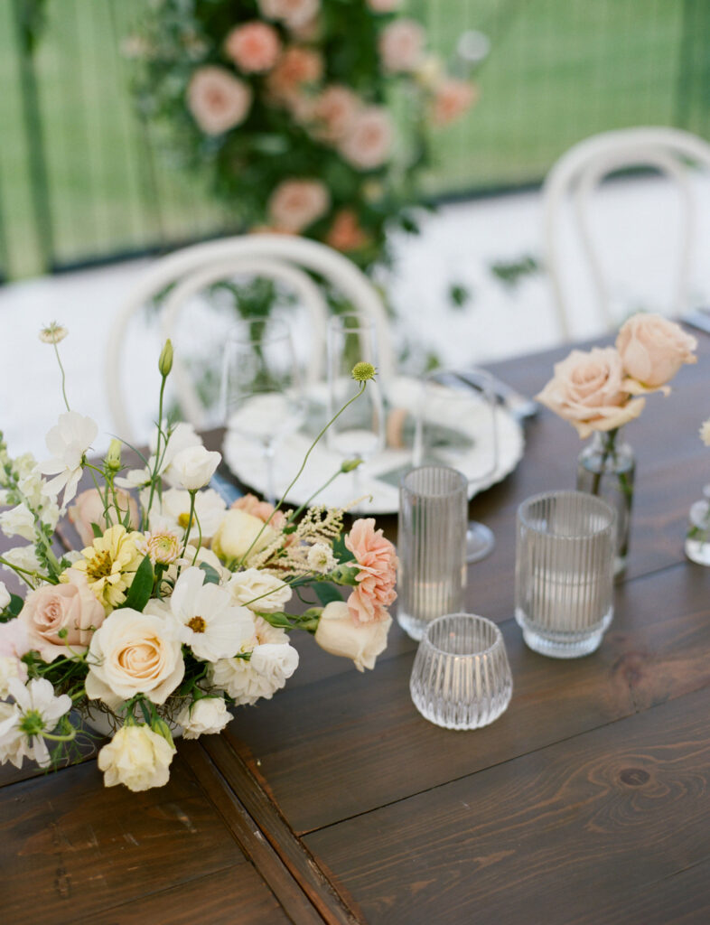 A wooden table is decorated with pastel flower arrangements, candles in glass holders, and elegant tableware. A floral backdrop is visible in the background.