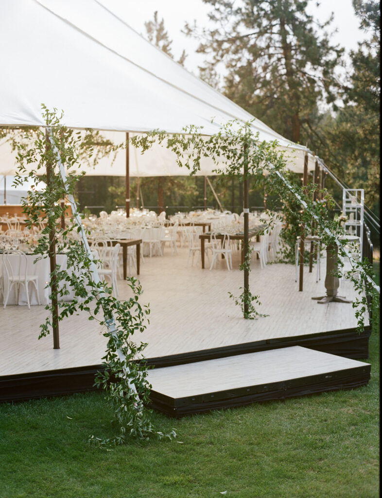 Outdoor wedding venue with a large white tent, decorated with green vines. Inside are white tables and chairs arranged neatly. Green lawn surrounds the setup under tall trees.