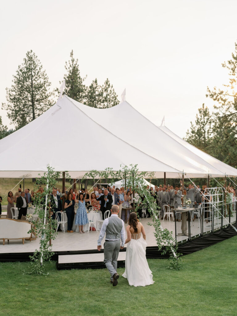 A couple walks toward a large white tent with seated and standing guests inside. The setting is outdoors with trees in the background.