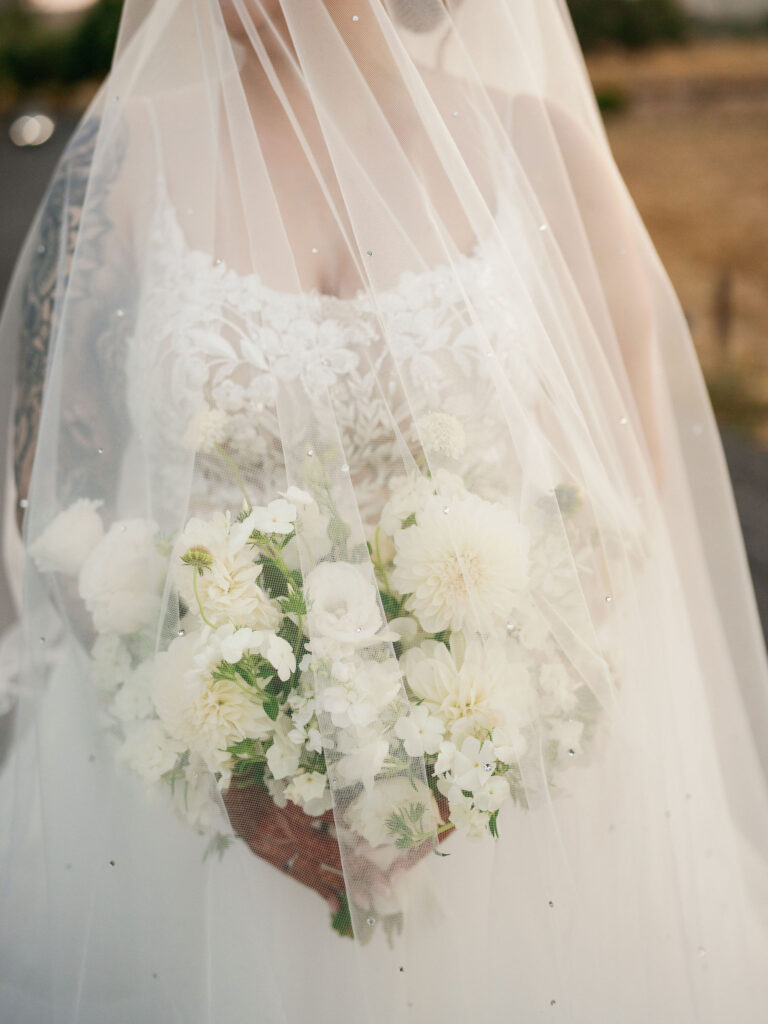 A bride holds a bouquet of white flowers, partially covered by a sheer veil. Her dress features lace details and there's a visible tattoo on her arm.