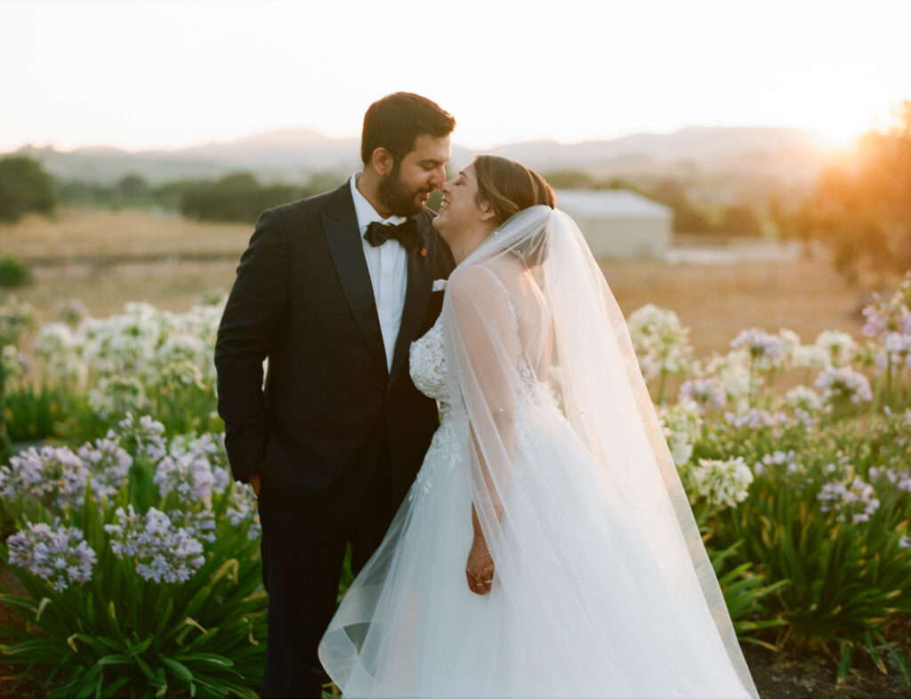 A bride and groom stand smiling at each other in a breathtaking outdoor setting, expertly crafted by Carneros Wedding Planner, surrounded by vibrant flowers and bathed in the warm glow of a sunset.
