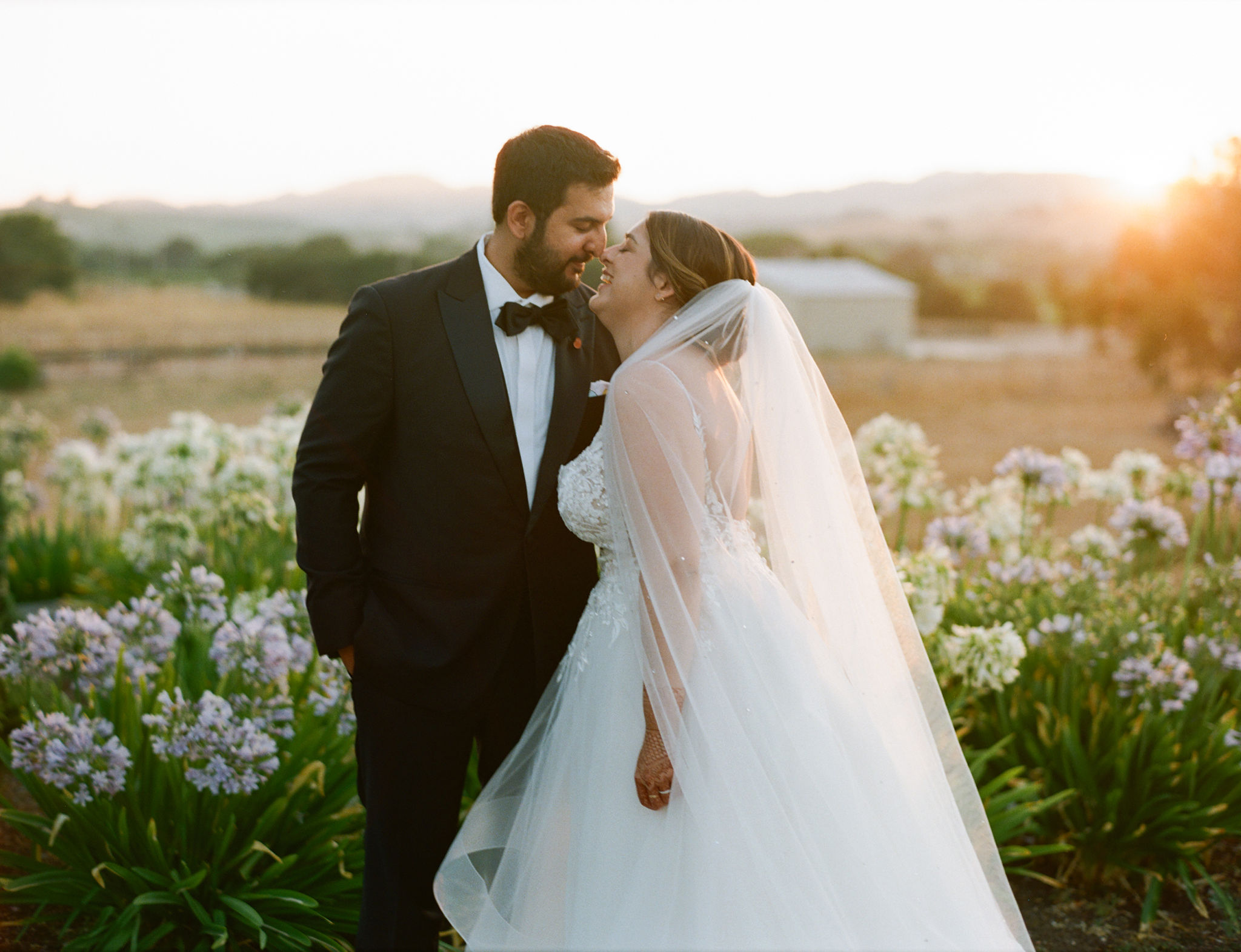 A bride and groom stand smiling at each other in a breathtaking outdoor setting, expertly crafted by Carneros Wedding Planner, surrounded by vibrant flowers and bathed in the warm glow of a sunset.