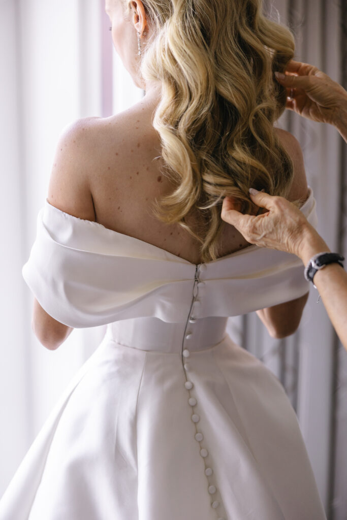 A woman in an off-the-shoulder wedding dress is having her hair adjusted.