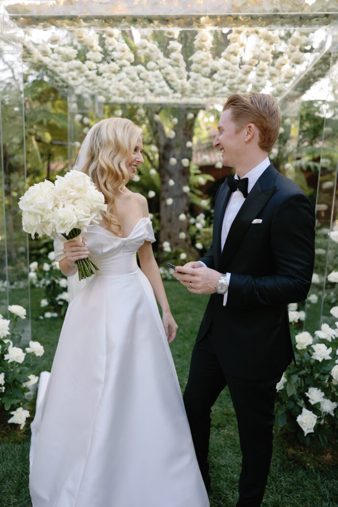 A bride in a white dress holds a bouquet of white roses, standing beside a groom in a black suit with a bow tie, under a canopy decorated with white flowers. They are smiling at each other.