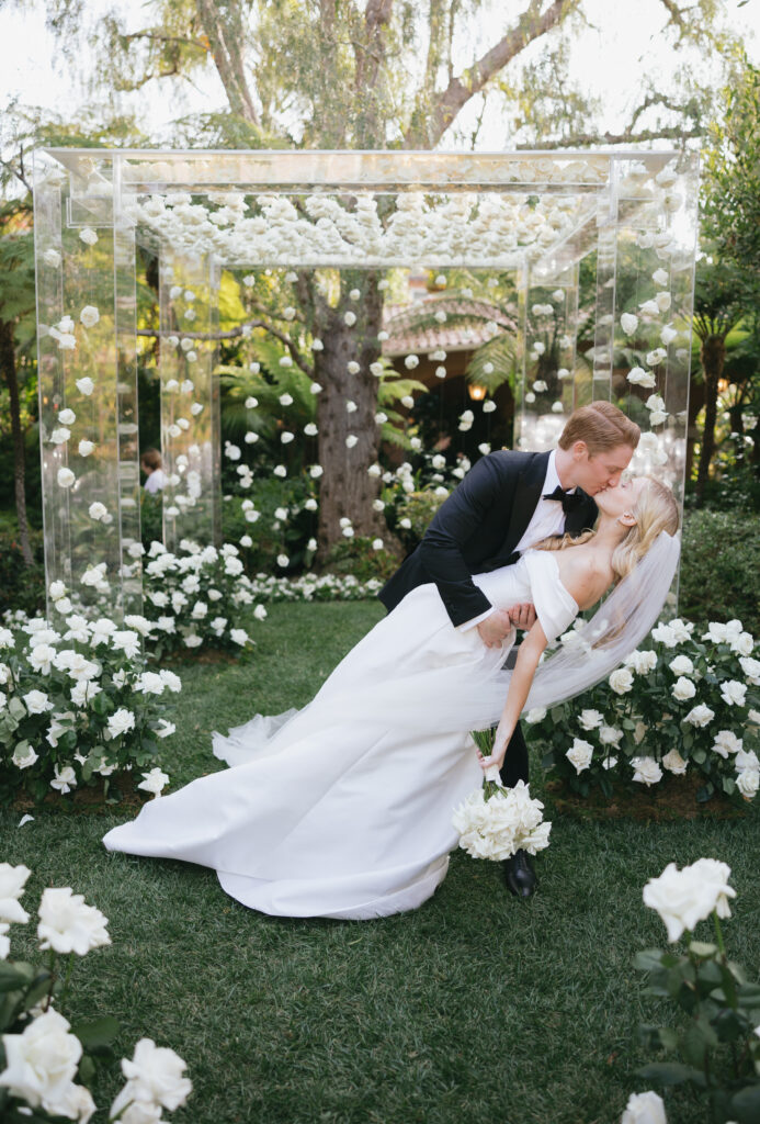 A couple kisses under a clear floral arch surrounded by white roses in a garden setting.