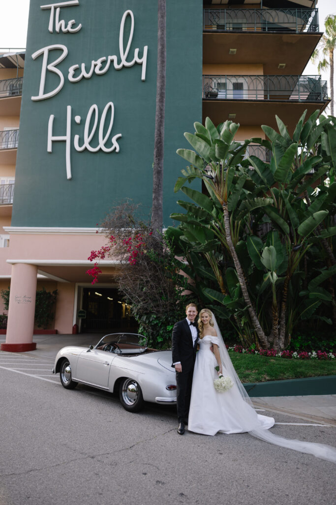 A bride and groom pose in front of a silver car and the Beverly Hills Hotel, with tropical plants and the hotel's sign in the background.