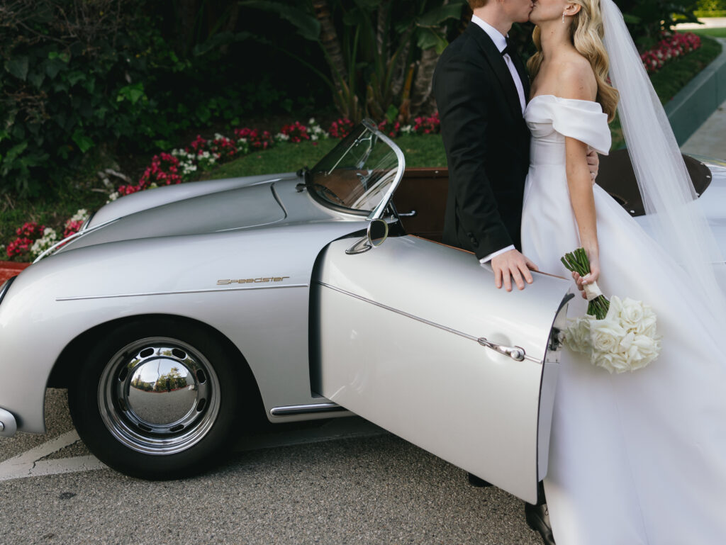 Bride and groom kissing beside a vintage silver car. Bride holds a bouquet of white roses.