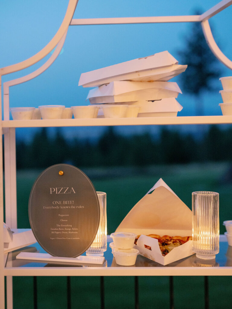 A shelf displays pizza boxes, small sauce containers, and lit candles. A nearby sign reads "Pizza: One Bite! Everybody knows the rules." The background shows a grassy field.