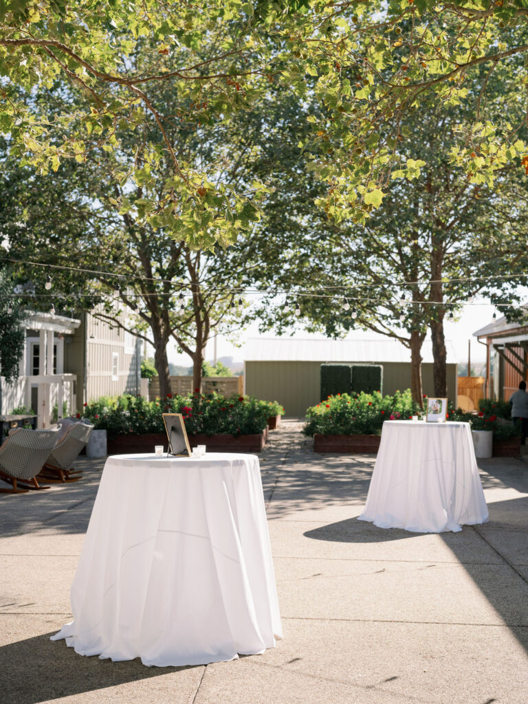 Outdoor space with two round tables draped in white cloth under tall trees. Each table holds framed photos. Buildings are visible in the background, and string lights hang above.