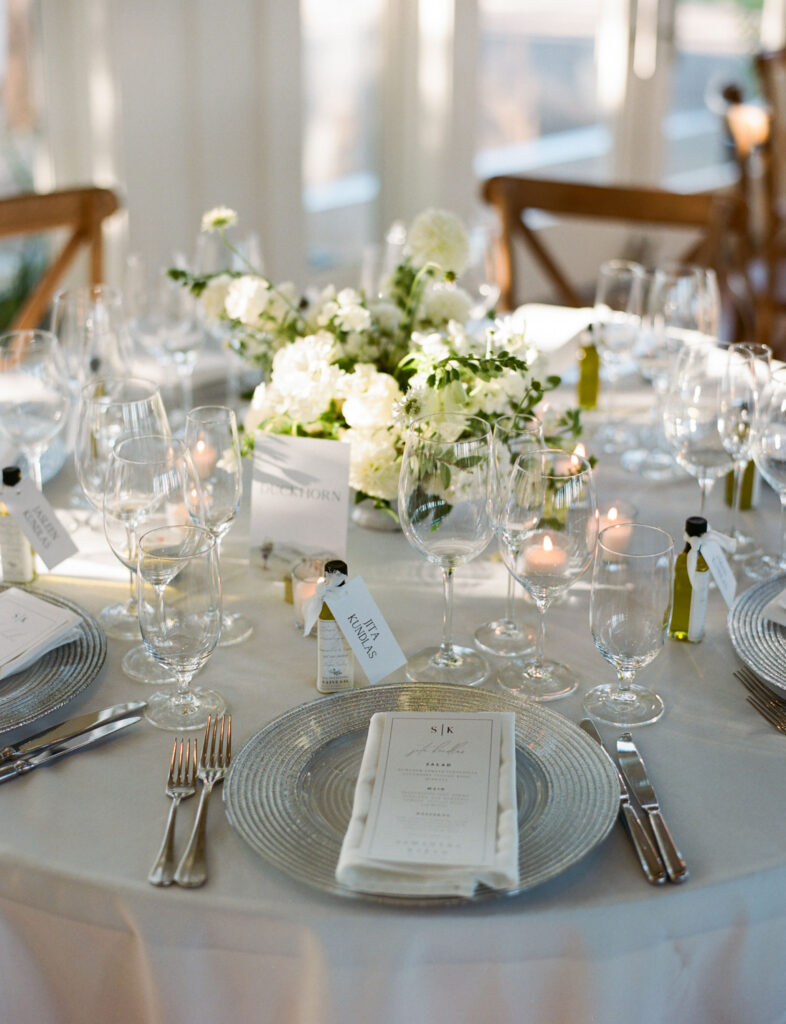 Elegant table setting with a white floral centerpiece, wine glasses, silverware, and a menu placed on a glass plate at a formal event.