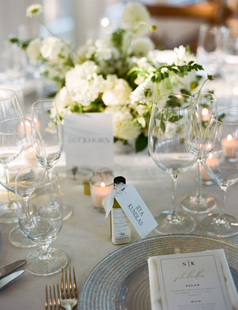 Elegant table setting with wine glasses, a floral centerpiece, and a small bottle with a tag. A menu is placed on a textured plate.
