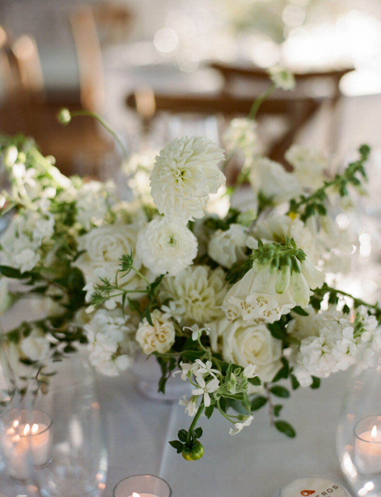 A floral arrangement with white roses, daisies, and greenery in a vase, surrounded by small lit candles on a table.