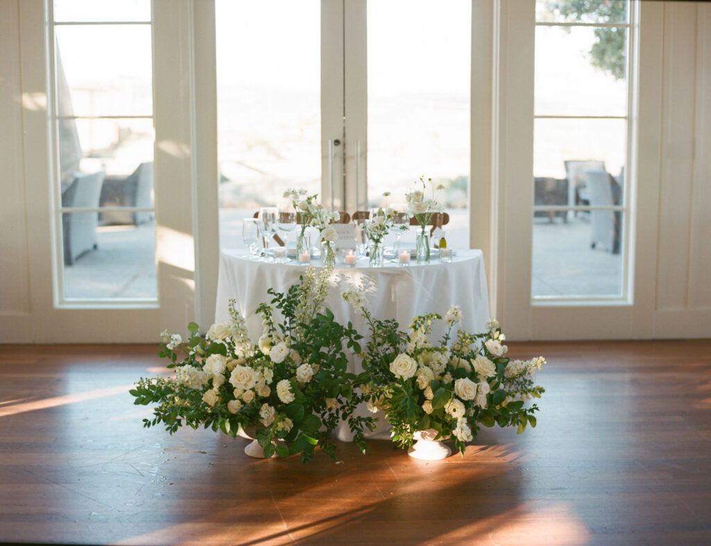 A round table decorated with white flowers and greenery is set for an event in front of large glass doors, with soft natural light streaming in.