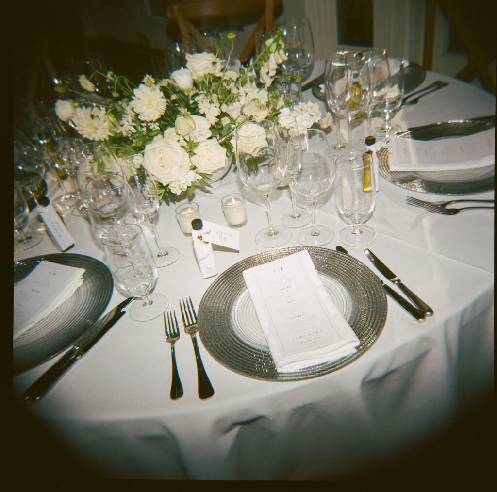 Elegant table setting with white floral centerpiece, glassware, cutlery, and menus on plates.