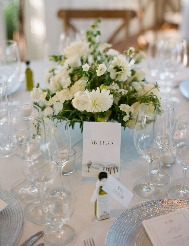 Wedding table setting with a floral centerpiece, a card labeled "Artesa," glassware, and a place card for Will Taylor on a small olive oil bottle.