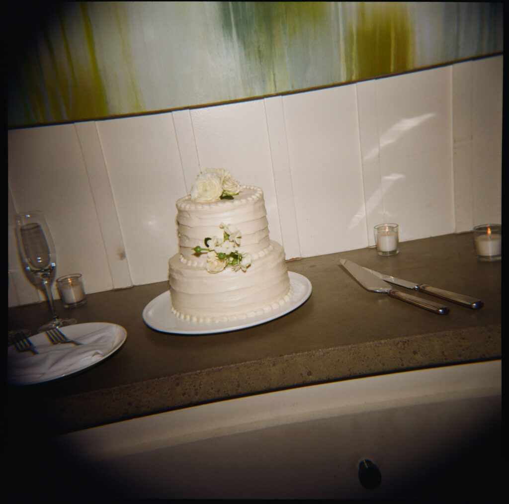 Two-tier white cake with flower decorations on a table, surrounded by a knife, fork, candles, and a glass.