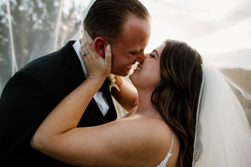 A bride and groom share a close, joyful moment under a veil, smiling at each other.