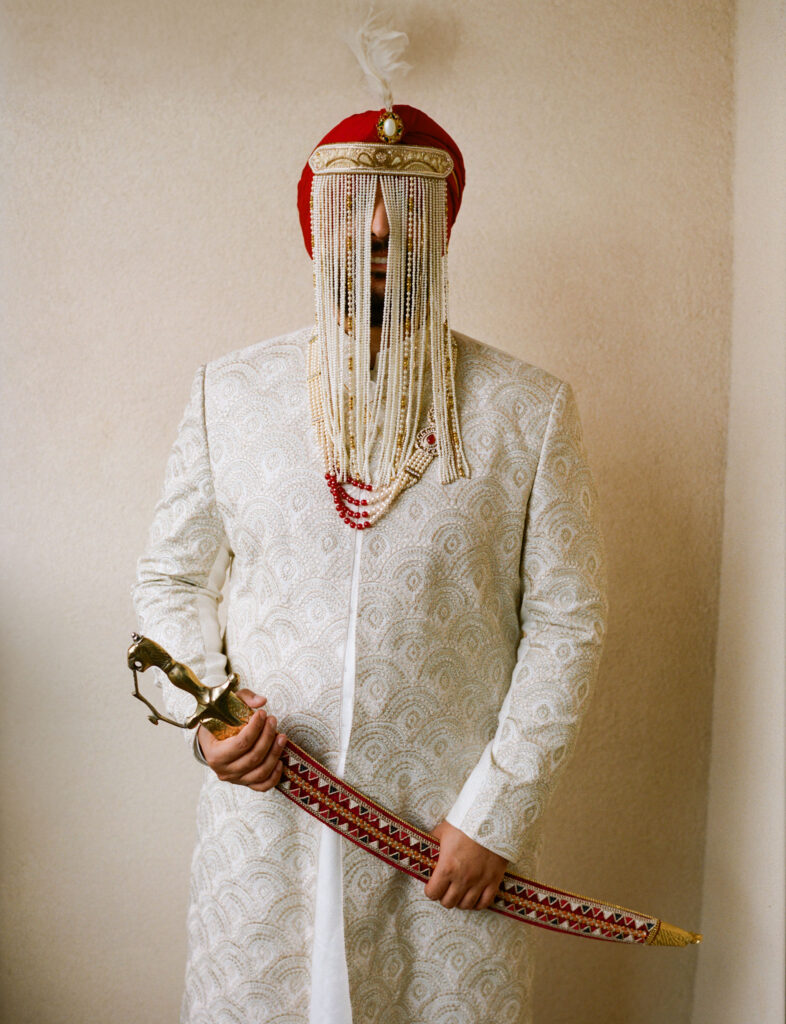 A person in ornate attire, wearing a beaded headdress with a face veil, holds a decorative sword.