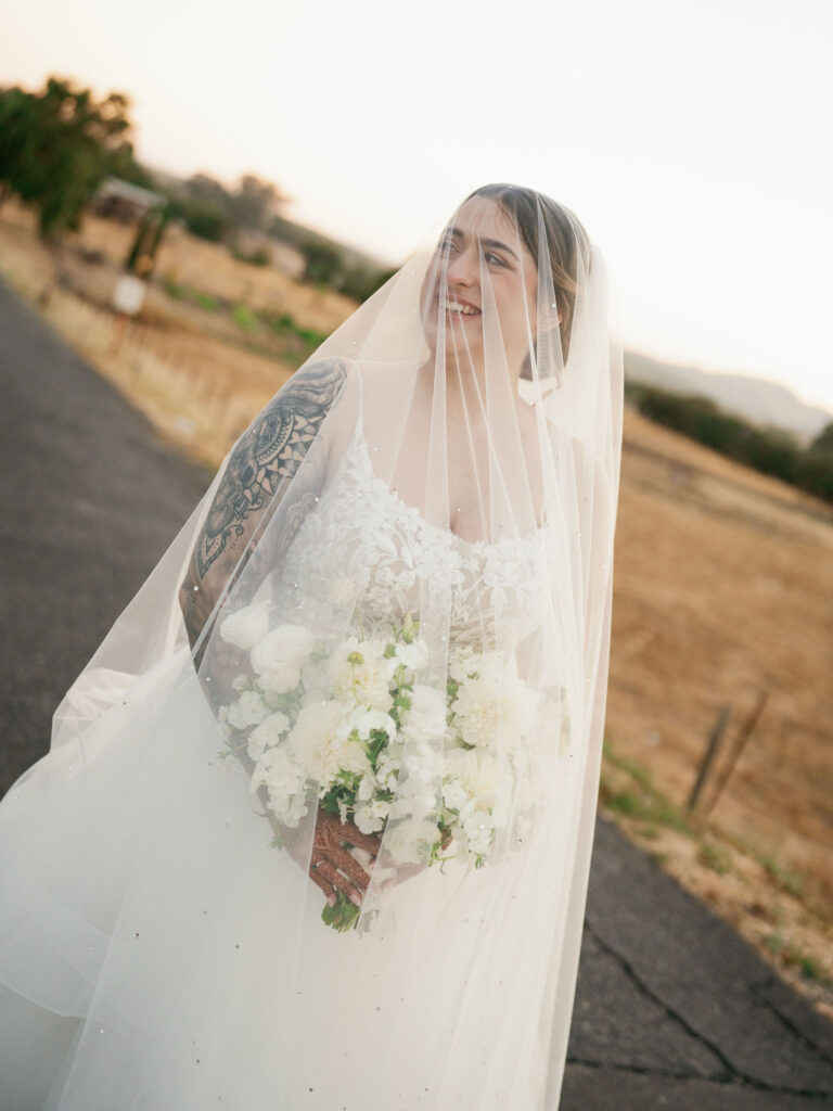Bride in a white dress holding a bouquet of white flowers, standing on a rural road at sunset, with a veil covering her face and shoulders.