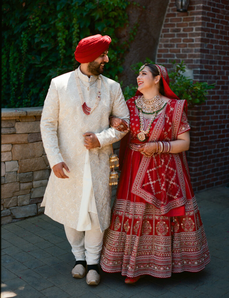 A couple in traditional Indian attire pose outdoors; the man wears a white sherwani and red turban, while the woman wears a red lehenga and ornate jewelry.