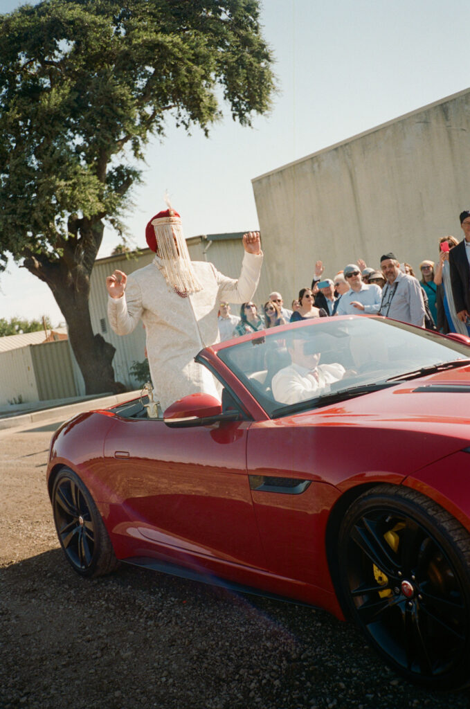 A person wearing festive attire stands in a red convertible during a celebration, with a crowd and a tree in the background.