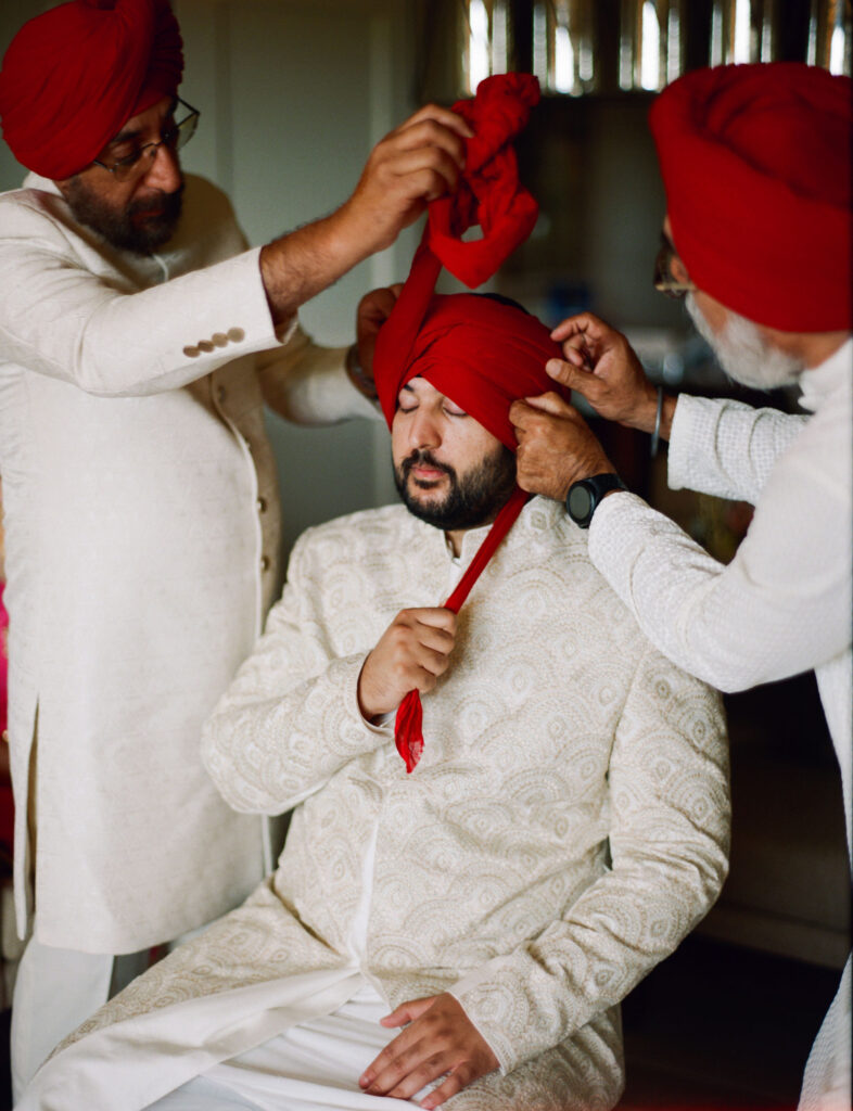 Three men are tying a red turban on a seated man's head, who is wearing a cream-colored embroidered outfit.