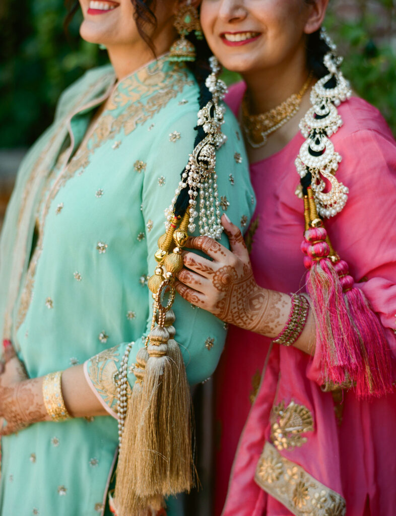 Two women in traditional attire with intricate embroidery and jewelry, displaying henna designs on their hands.