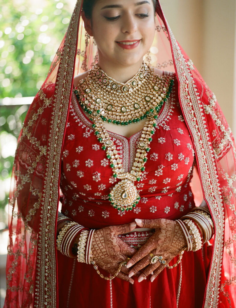 A person in a red traditional outfit with intricate jewelry and henna on hands, smiling with eyes closed.