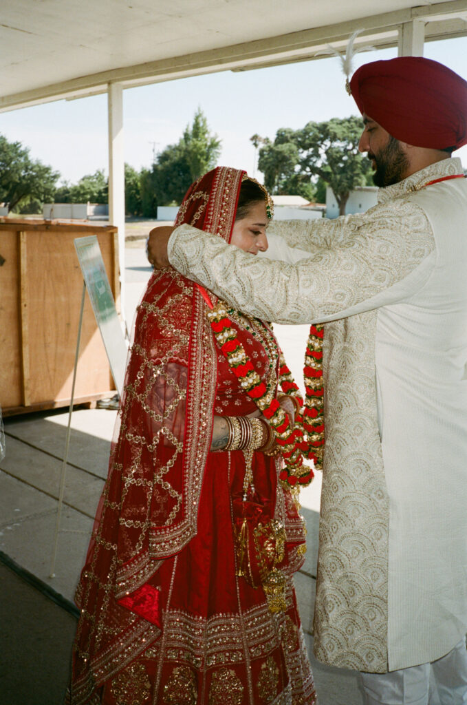 A man and woman in traditional Indian attire exchange garlands in an outdoor setting.