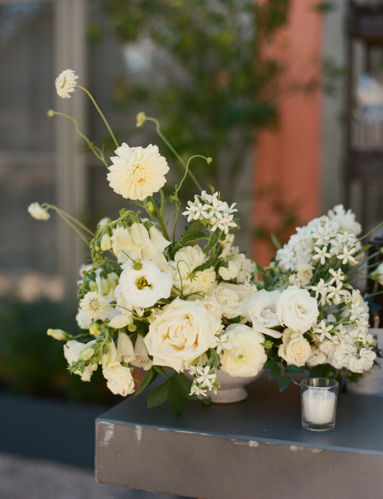 A floral arrangement of white roses and assorted flowers in a vase on a table, with a small candle beside it.