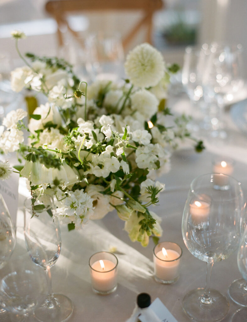 A white floral centerpiece with green accents on a table, surrounded by lit votive candles and clear wine glasses.