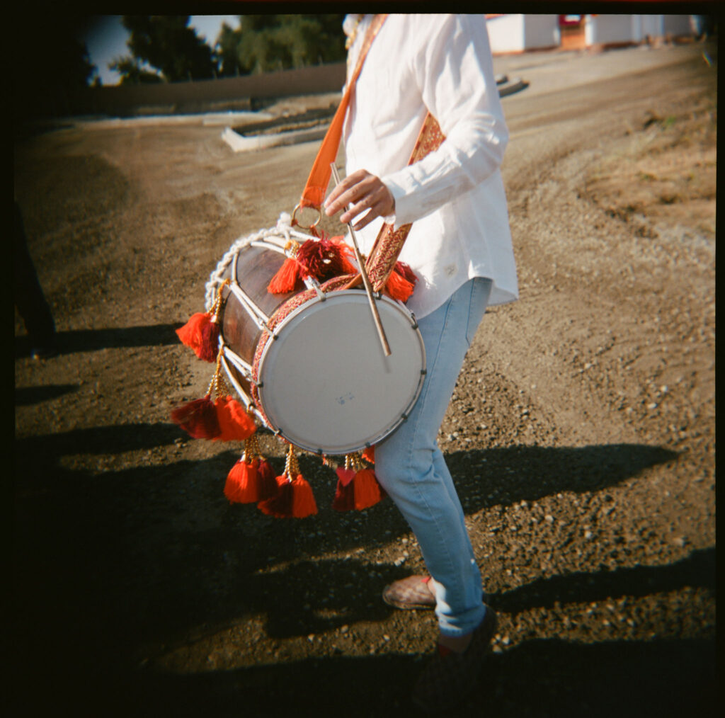 Person in a white shirt and jeans playing a drum adorned with red tassels outdoors.