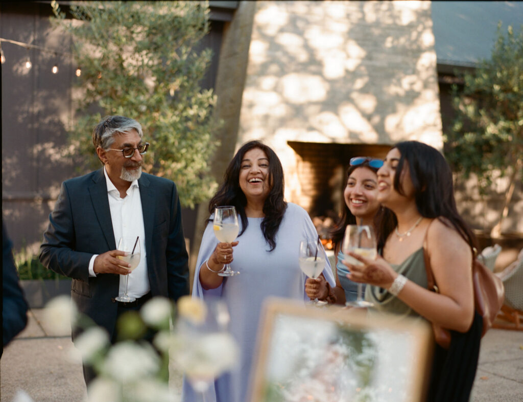 A group of people smiling and holding drinks while standing outdoors near a fireplace.