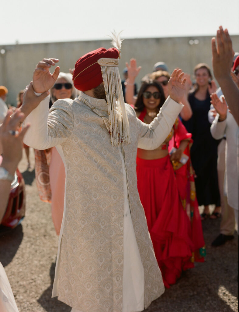 A person in a decorative outfit and turban adorned with strings is dancing amid a group of people outdoors.