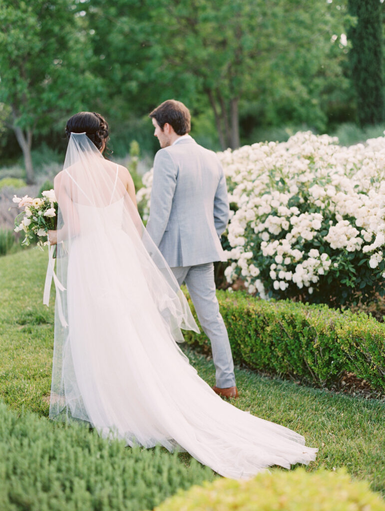 A bride and groom walk through a garden, with the bride in a white dress and veil and the groom in a light gray suit. They are surrounded by blooming white flowers and greenery.