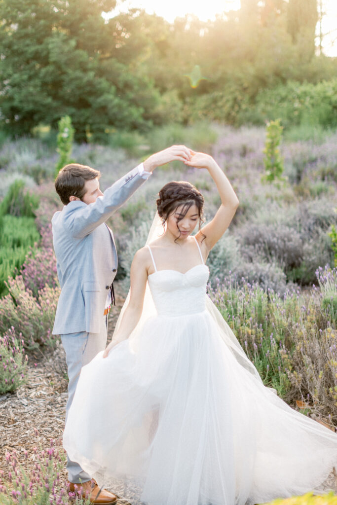 A couple dances in a garden with blooming flowers. The woman wears a white wedding dress, and the man is in a light gray suit. Sunlight filters through the trees.