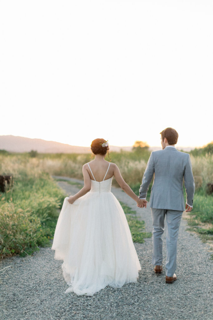 A couple holding hands walks down a gravel path during sunset. The woman wears a white dress, and the man wears a light gray suit.