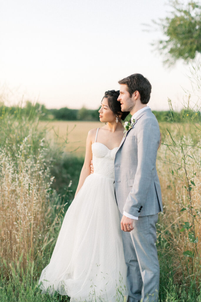 A couple in wedding attire stands in a grassy field, gazing into the distance under a pale sky.