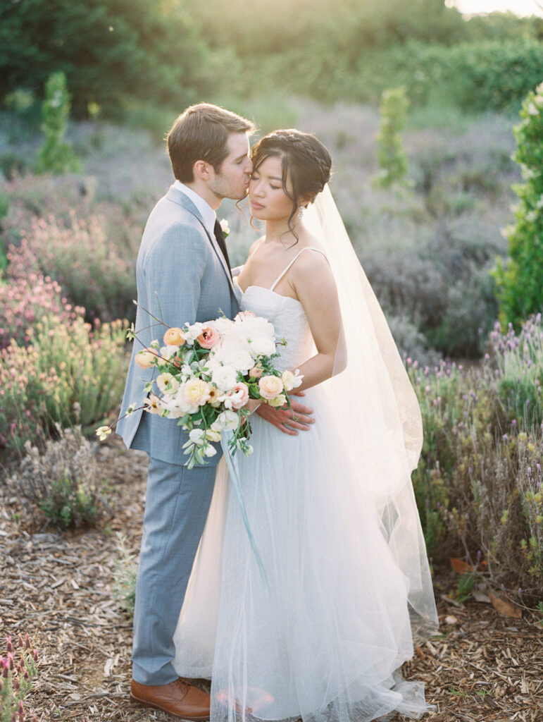 Bride and groom stand in a garden, with him in a light suit kissing her forehead. She holds a bouquet of white and pink flowers, wearing a veil and white gown.