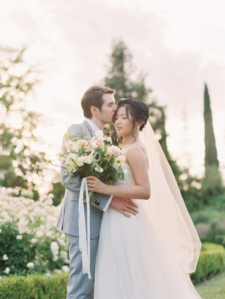 A couple in wedding attire embraces in a garden. The groom kisses the bride's forehead while she holds a bouquet. The setting is soft and romantic with trees in the background.