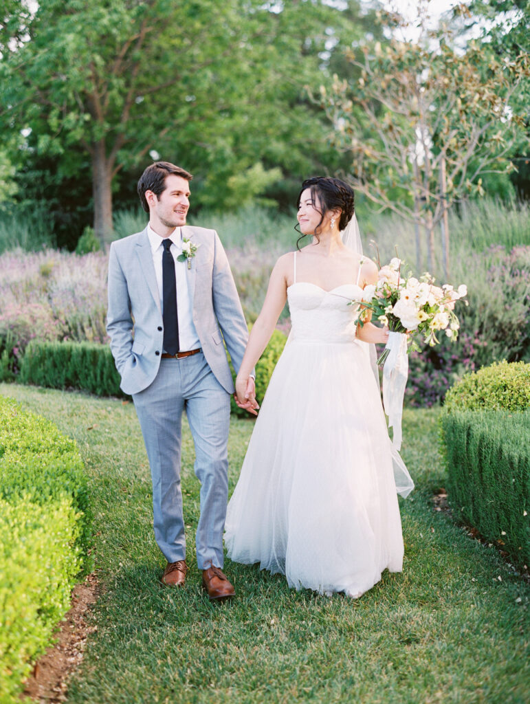 A bride and groom walk hand in hand down a garden path. The bride holds a bouquet, and both smile at each other.