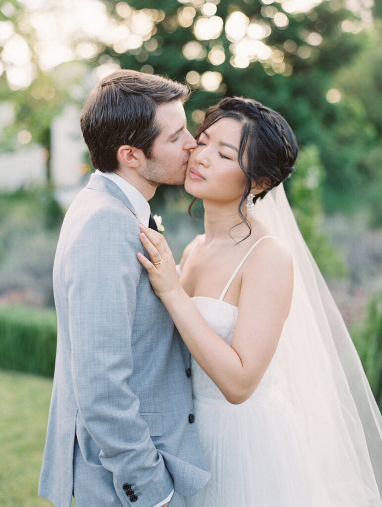 A groom in a light gray suit kisses the cheek of a bride in a white dress and veil, standing outdoors with greenery in the background.