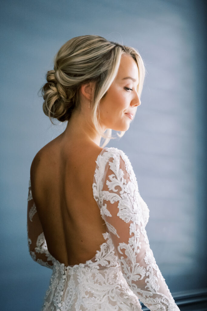 Bride with blonde hair in an elegant updo wearing a lace wedding dress with an open back stands against a soft blue background, looking to the side.