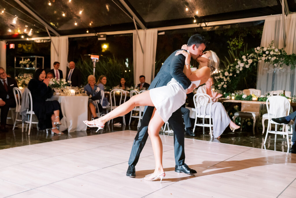 A couple in formal attire perform a dance at a wedding reception, with the woman lifted and dipped by the man. Guests are seated at tables in the background under a tent with string lights.