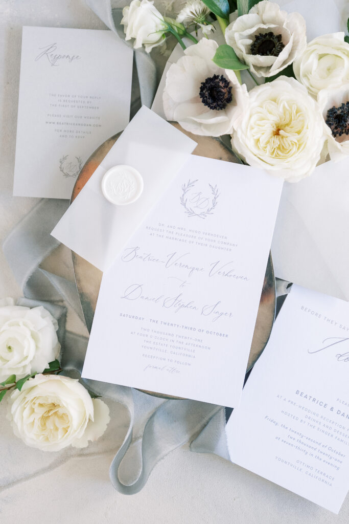 Elegant wedding invitation suite with floral designs, surrounded by white roses and greenery on a soft textured background.