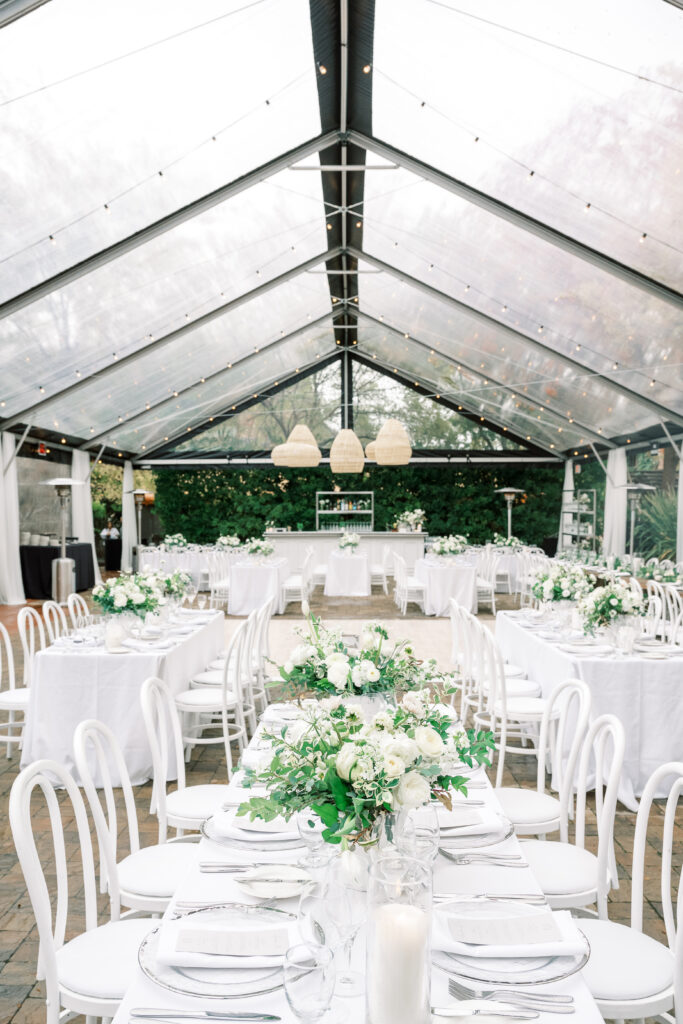 Elegant outdoor wedding reception setup under a clear tent, featuring white tables and chairs with green and white floral centerpieces. Hanging pendant lights create a warm atmosphere.