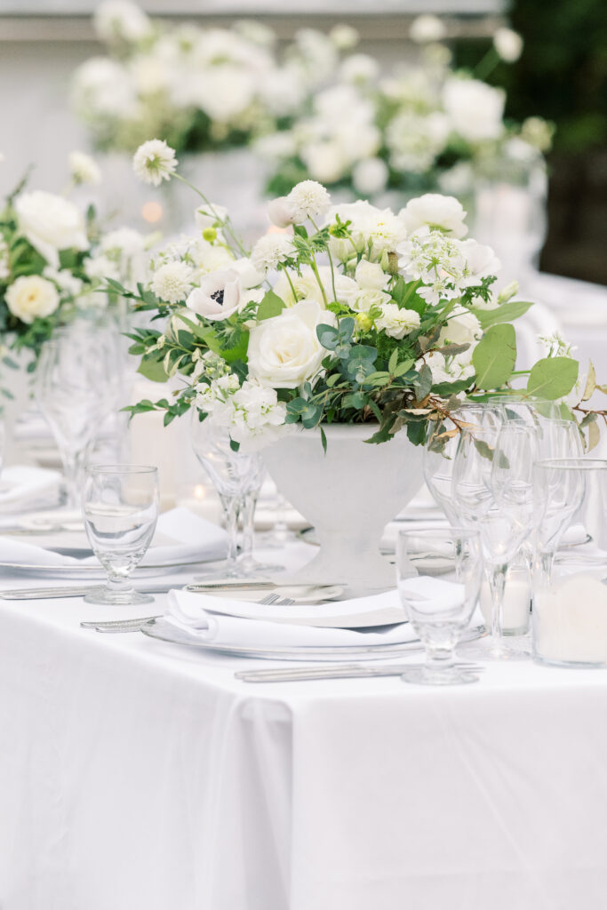A white tablecloth-covered table is elegantly set with white floral centerpieces, glassware, and neatly folded napkins.